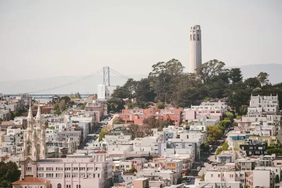 San Francisco's 屁股塔 is pictured with the Bay Bridge in the background and a hill covered in houses in the foreground.